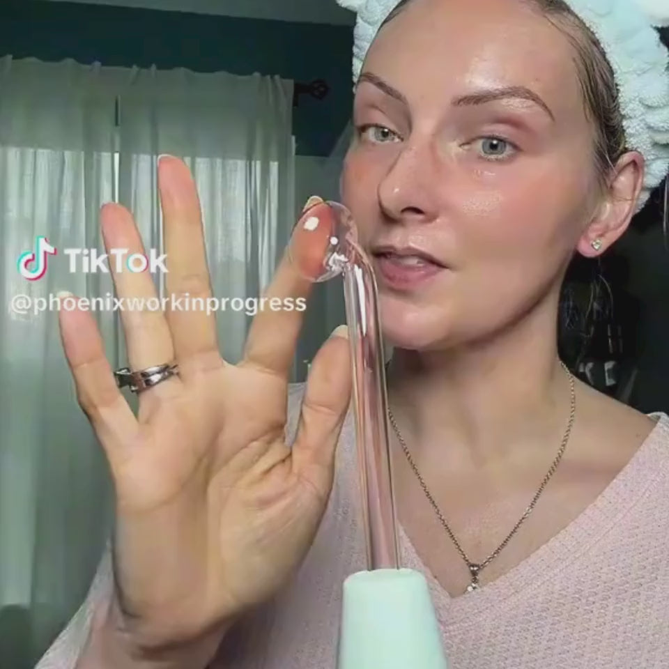 A woman with a white towel wrapped around her head and bare, freshly cleansed skin films a TikTok skincare review (@phoenixworkinprogress). She holds up the white "Glow Anti-Aging Facial Conductive Gel" pump bottle and talks through the product while gesturing with her free hand. She wears a pink ribbed top and a delicate necklace, filmed in a bright room with sheer curtains in the background. A TikTok watermark and her username appear on screen